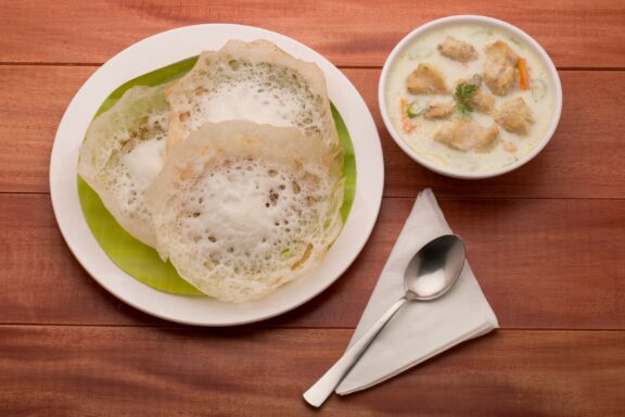 A plate of appam with a side of chicken stew, served on a wooden table.