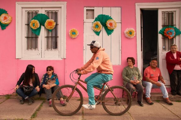 A cyclist rides past a pink building with green windows and paper flowers in Chile, observed by onlookers.