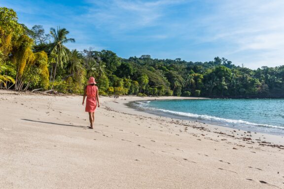 A person in a red dress walking on a sandy beach in Costa Rica with lush greenery in the background and a clear blue sky above.