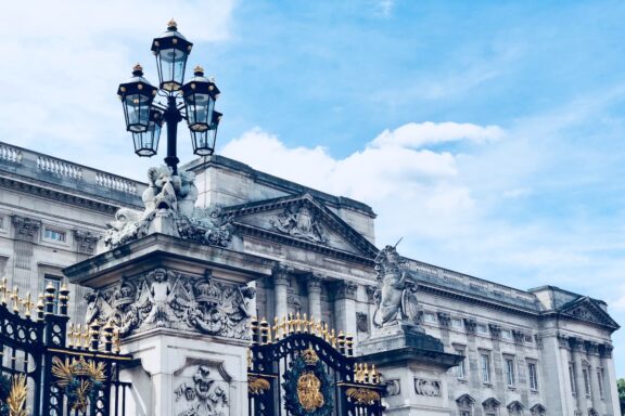 An ornate street lamp in front of a grand, classically designed building with sculptures, under a partly cloudy sky.