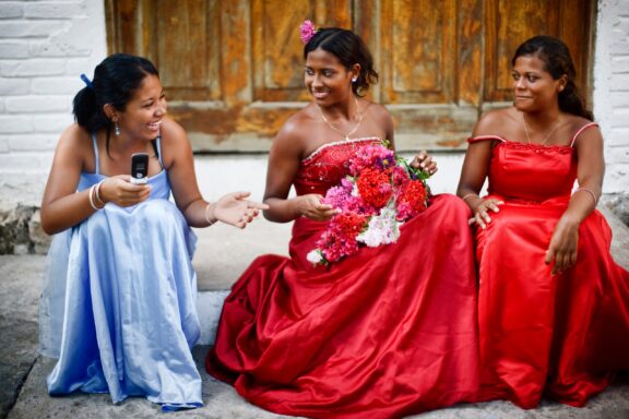 Three girls in El Salvador, two in red dresses and one in blue, are chatting and smiling near an old wooden door.