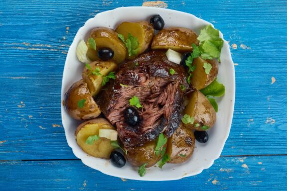 Greek lamb kleftiko dish with roasted lamb, potatoes, olives, and herbs, served on a white plate with a blue background.