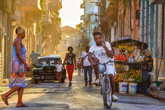 A street scene in Havana, Cuba with people walking and a person riding a bicycle, vintage cars, and a fruit stand on a sunny day.