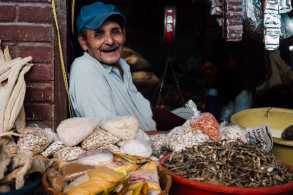 A smiling shopkeeper in Honduras sitting beside various dry goods including beans and grains, with a Coca-Cola sign visible in the background.