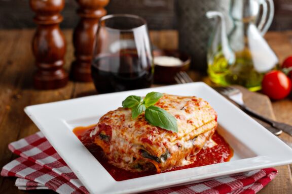 A lasagne slice with a basil leaf, served with red wine, olive oil, and tomatoes, implies an Italian meal setting.