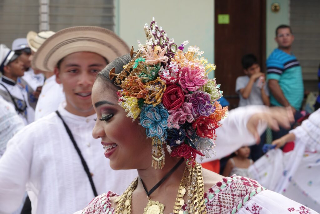 A woman in Panamanian attire and floral headdress smiles at a cultural event in Los Santos, Panama, with a man in white behind her.