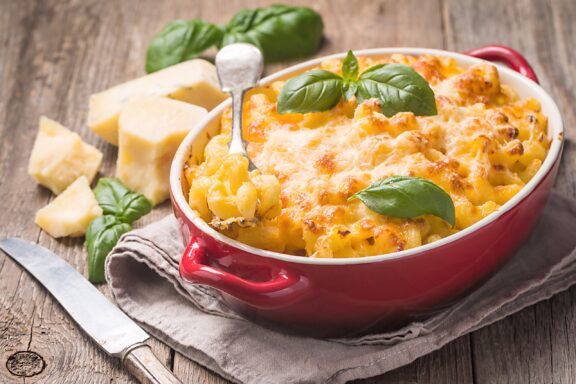 Baked macaroni and cheese with a melted cheese topping and basil garnish, served in a red dish with cheese pieces and a knife nearby.