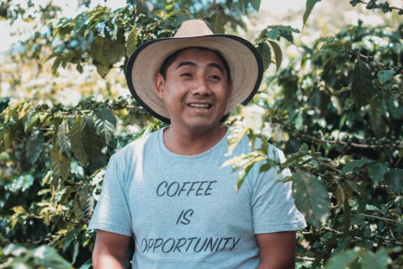 A man wearing a hat and a t-shirt that reads "COFFEE IS OPPORTUNITY" is smiling and standing in front of green foliage, possibly in Antigua.