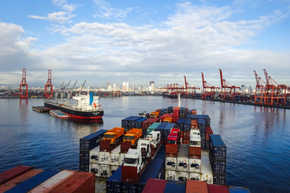 A cargo ship docked at the Port of Manila, Philippines, with containers stacked on the vessel and on the dock, under a partly cloudy sky.