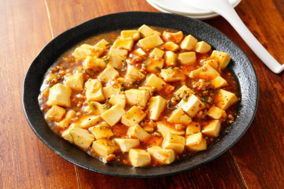 A dish of Mapo Tofu, a traditional Chinese dish, featuring cubed tofu in a spicy sauce with minced meat, served in a black bowl on a wooden table.