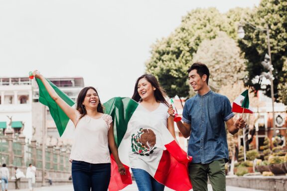 Three people smiling and walking outdoors, two of them holding a Mexican flag, with trees and a building in the background.