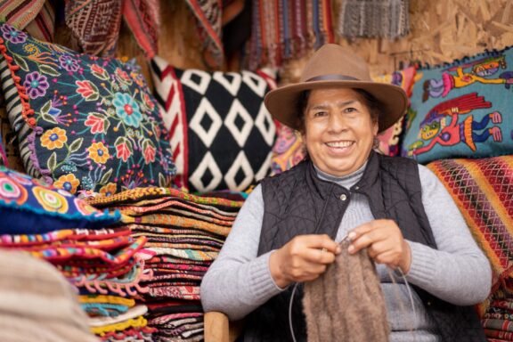 A smiling Peruvian woman wearing a hat and a vest, sitting in front of colorful textiles, possibly at a market in Peru.