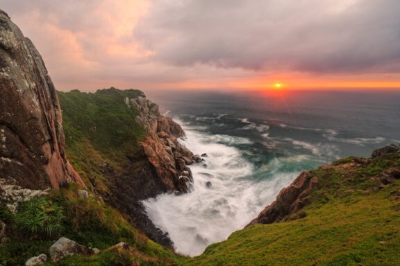 A scenic view of Praia do Rosa with a sunset over the ocean, waves crashing against rocky cliffs, and lush greenery on the coastline.