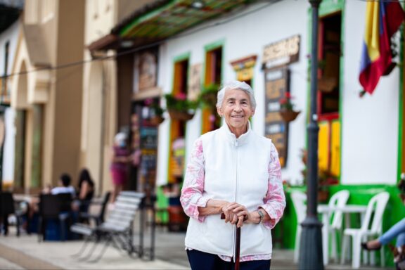 A smiling elderly woman with a cane standing on a sidewalk in Salento, Colombia, with colorful buildings and a café terrace in the background.