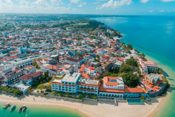 Aerial shot of Stone Town, Zanzibar displays dense historical architecture, red roofs, clear blue coastline, and boats.