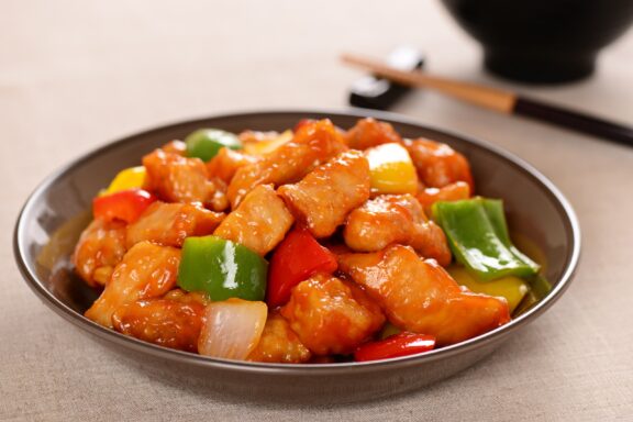 A dish of sweet and sour pork with chunks of pineapple, bell peppers, and onions, served on a plate with chopsticks in the background.