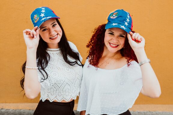 Two smiling women wearing hats with the Venezuelan flag, standing against an orange wall.