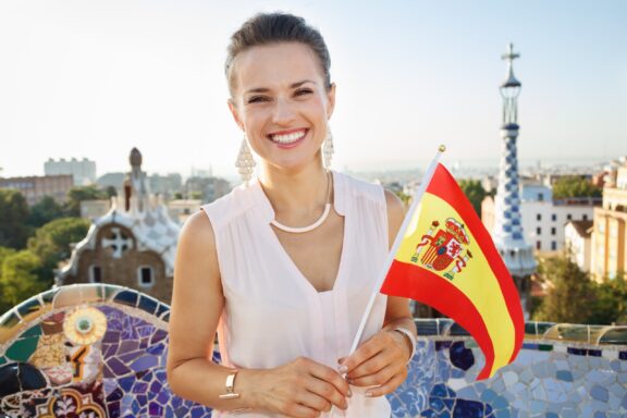 A smiling woman holding a Spanish flag with a scenic city view in the background, possibly in Spain.