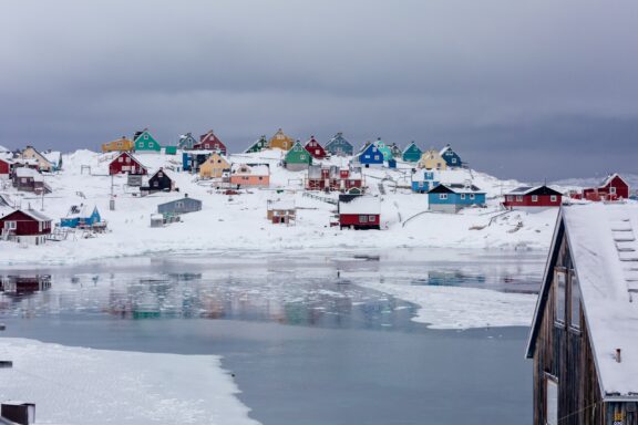 A view of Aasiaat, Greenland, featuring colorful houses scattered on snowy terrain with ice floating on the water in the foreground.