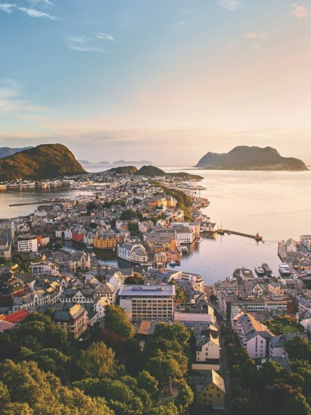 Aerial view of Ålesund, Norway shows dense architecture on a peninsula, encircled by water and mountains at twilight.