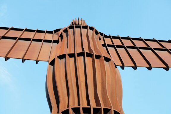 An image of the Angel of the North, a large steel sculpture of an angel with outstretched wings, set against a clear blue sky.