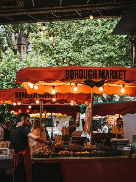 A lively scene at London's Borough Market with orange-canopied stalls, diverse goods, browsing customers and a foreground vendor.