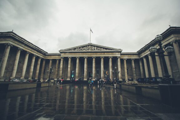 A wide-angle view of the British Museum in London on an overcast day, with visitors walking in front of the building's grand columned facade.