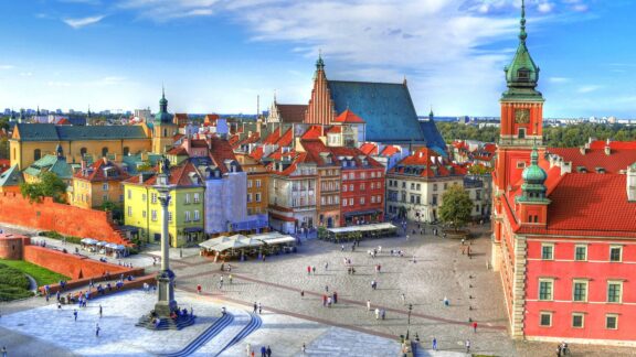 A panoramic view of Castle Square, featuring colorful buildings, a large open square with people, and a prominent statue, under a partly cloudy sky.