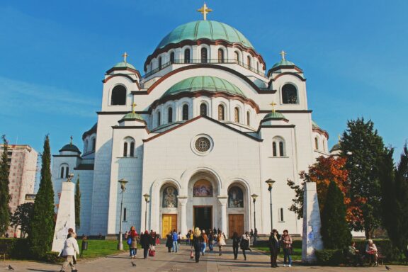 The photo showcases the large white Church of Saint Sava in Belgrade, Serbia, with people strolling in front.