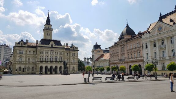 City Hall in Novi Sad, Serbia, with people sitting on benches and walking in a square, historic European architecture under a partly cloudy sky.