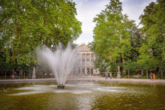 A fountain in a pond, with green trees and a classic building, possibly in Brussels Park, with people enjoying the scene.