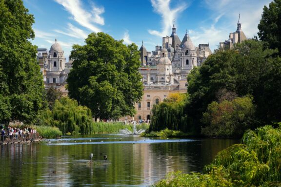 A picturesque view of Hyde Park, London, showcasing a lake, trees, a historic building, under a semi-cloudy sky.