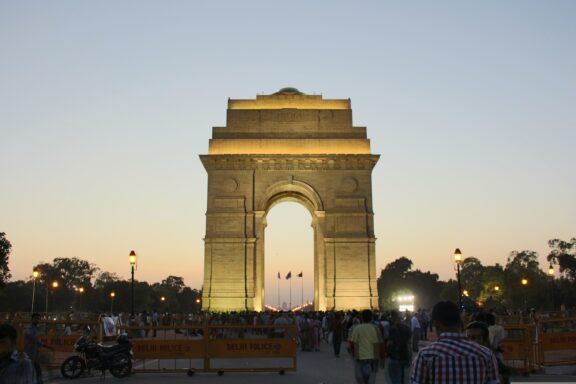 India Gate monument at dusk with people milling around and lights beginning to illuminate the area.