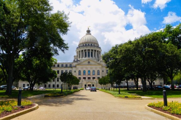 Mississippi State Capitol in Jackson is pictured with a clear sky, green trees, and a pathway leading to it.