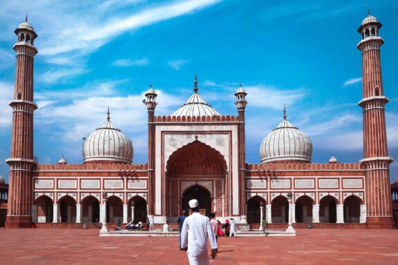 A person walking towards the entrance of Jama Masjid, a large mosque with two tall minarets and three domes under a blue sky with wispy clouds.