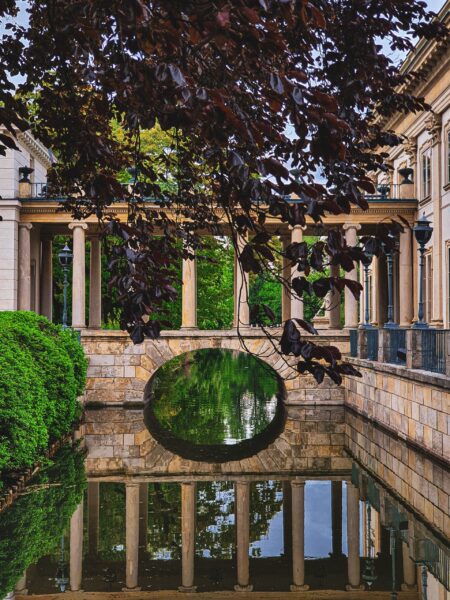 A serene view of Łazienki Park with a classical architecture building, a calm pond reflecting the structure, and lush greenery surrounding the area.