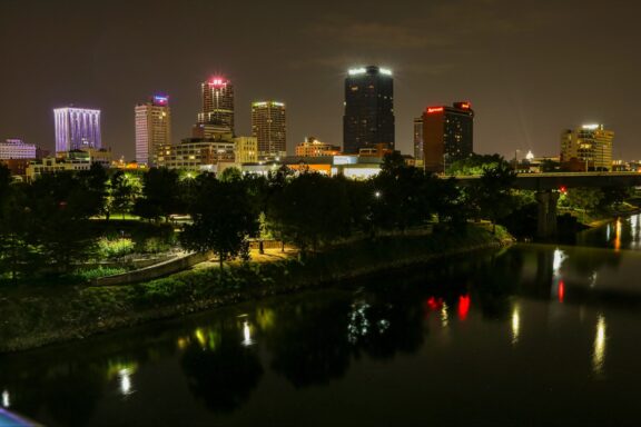 A nighttime cityscape of Little Rock, Arkansas, with illuminated buildings reflected in a body of water.