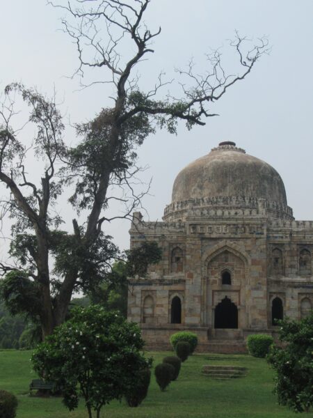 A historical dome structure with intricate carvings at Lodi Gardens, surrounded by trees and greenery, under a hazy sky.