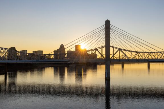 Sunset view of the Louisville Bridge in Kentucky with the sun casting a warm glow on the skyline and reflecting on the water's surface.