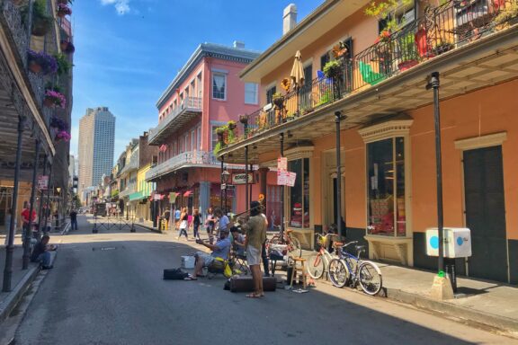 A vibrant New Orleans street scene with colorful buildings, plant-filled balconies, pedestrians, and parked bicycles.