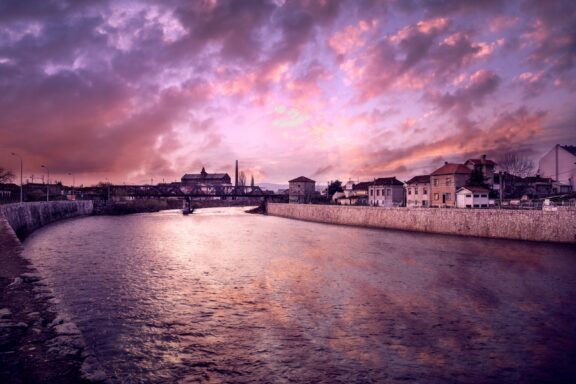 A scenic view of a river at dusk with a colorful sky in Niš, Serbia, featuring a bridge in the distance and buildings along the riverbank.