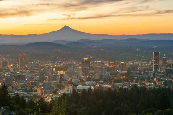 A dusk view of Portland, Oregon with city lights, Mount Hood, and an orange sky.