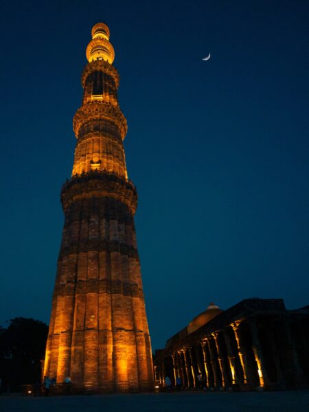 Illuminated Qutb Minar against a twilight sky with a crescent moon, located in Delhi, India.