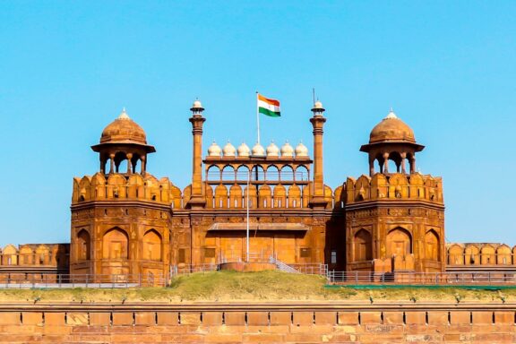 A photo depicts the Red Fort's red sandstone architecture, two domed towers, and the Indian flag under a clear blue sky in Delhi.