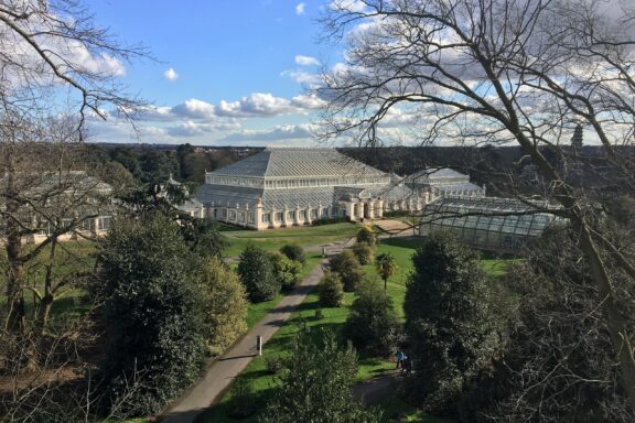 A large glass greenhouse is the centerpiece of the Royal Botanic Gardens at Kew, surrounded by trees under a partly cloudy sky.