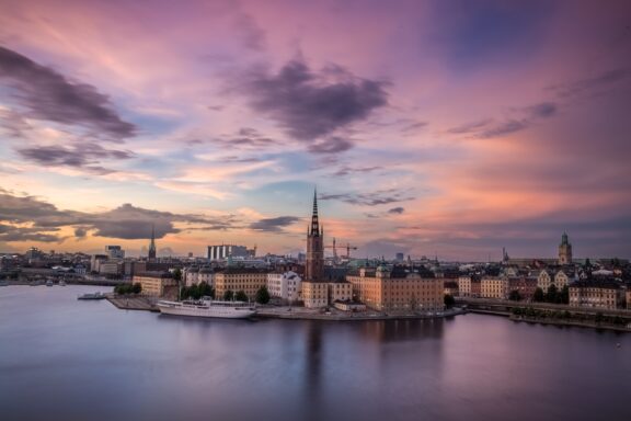 A sunset in Stockholm, Sweden displays pastel skies reflected in calm water, highlighting city architecture and a tower.