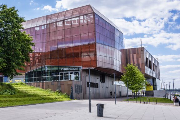 A modern "Planetarium" building, possibly part of the Copernicus Science Center Museum, with a glass and metal facade under a cloudy sky.