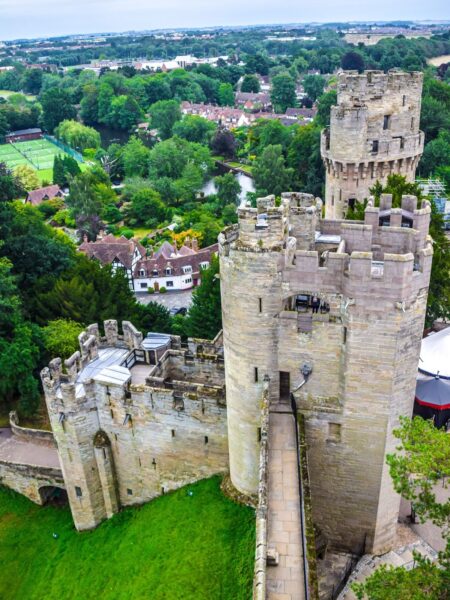 Aerial view of Warwick Castle with its prominent central tower, surrounded by green lawns and trees, with a backdrop of a residential area.