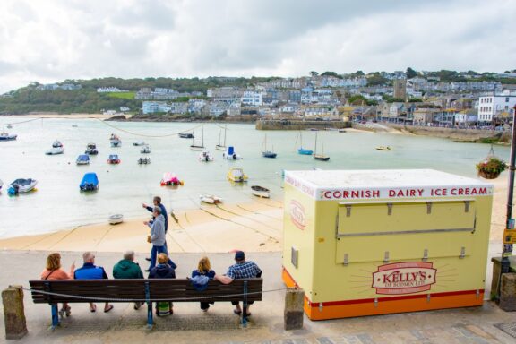 People sit on a bench overlooking a harbor, boats, a Cornish ice cream cart, and a coastal town on a hill.