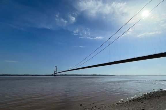 A photo from a riverbank showcases a suspension bridge's long cables and tower, with a wide river and clear blue sky behind.